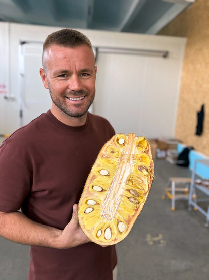 A Iulian Perseca holding a halved fresh jackfruit showing large yellow edible pods and white seeds.