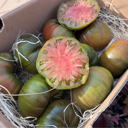 A box of ribbed green and red Marmande heirloom tomatoes on hay, with two halves showing a pink fleshy interior.