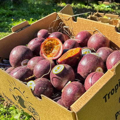 Fresh purple passion fruits in a cardboard box outdoors, with one sliced open to show the aromatic orange pulp and seeds.