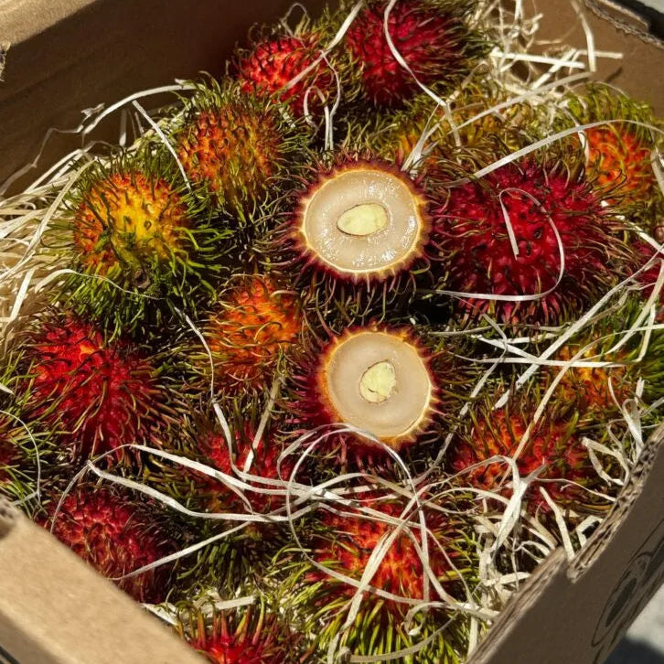A box of red Vietnamese rambutans on hay, with one fruit sliced open to show the translucent white flesh and center seed